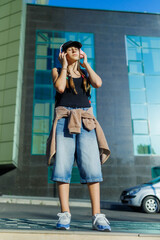 Fototapeta premium Gen Z, Relaxed teenager listening to music standing near a glass building.