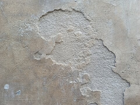 oA close-up shot of a weathered concrete wall with a large section of the outer plaster layer peeling and breaking away, exposing the rough and granular texture of the underlying cement beneath.