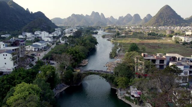 A stone arch bridge spans a calm river, framed by karst mountains and riverside settlements in a serene rural landscape.