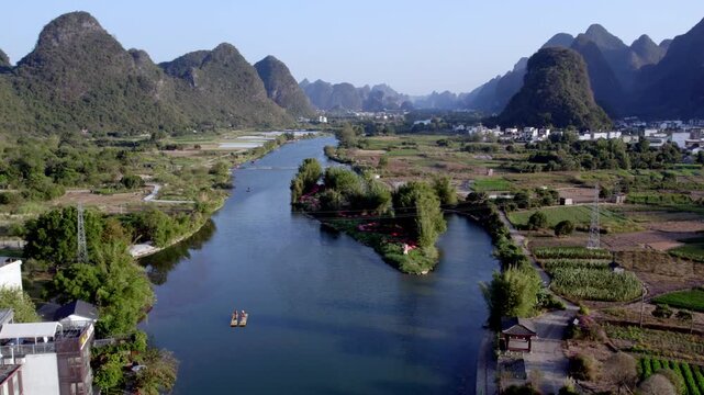 A river winds through a karst landscape, flanked by mountains, fields, and rural settlements with small boats on the water.