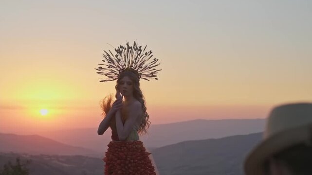 Tashkent, Uzbekistan - 4 11 2022: Film crew with a young female model in an autumn designer dress made of flowers and a headdress made of spikelets on the background of the sunset in the mountains.