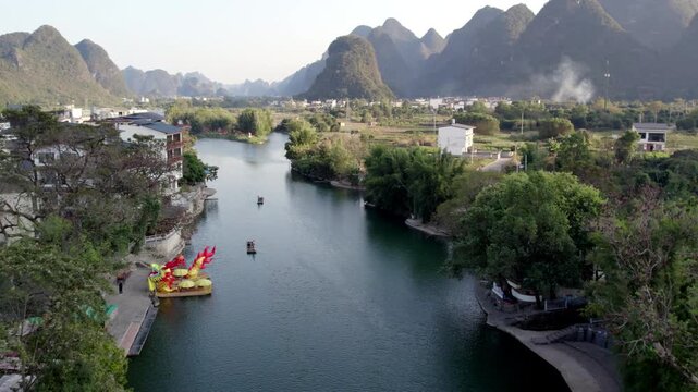 A stone arch bridge spans a calm river surrounded by karst mountains, with buildings, trees, and boats nearby.