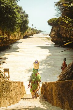 Zanzibar, Tanzania - 20th october, 2025: African women wearing traditional colorful kangas carrying yellow buckets balanced on heads walking down stone steps sandy cove Mtende Beach South Zanzibar
