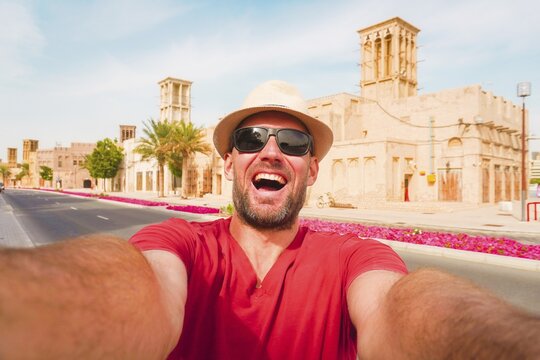 Happy male tourist wear red shirt and straw hat pose takes selfie at the Al Fahidi Historical Neighborhood in Old Dubai. Features a traditional wind tower backdrop cityscape view