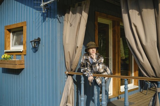 Pensive young woman leaning on railing of blue wooden cabin wearing hat and plaid shirt and looks thoughtful to distance. Bright sunlight shines on her as she enjoys warm weather outside