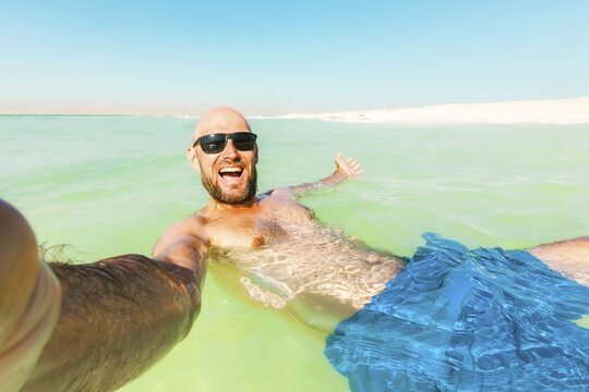 POV caucasian male Man floating wave pose send greetings in turquoise Dead Sea water wearing a traditional keffiyeh and sunglasses. Relaxing summer vacation at the lowest point on Earth in Jordan