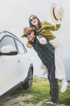 Portrait Happy couple Man with woman having fun piggyback next to white car on coastal cliff. Vast blue ocean and mountain horizon create romantic, joyful travel mood. Summer holiday adventure vibes