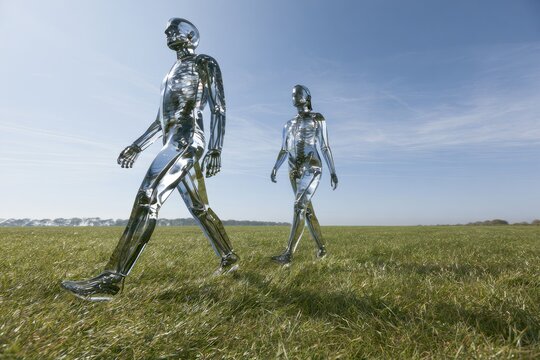 Two chrome robots walking across a grassy field under a blue sky.