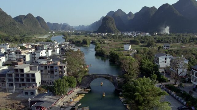 A rural riverside settlement with a stone arch bridge, surrounded by karst mountains and lush green landscapes.