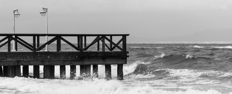 Black and white image of a wooden pier or boardwalk with rough sea waves crashing against its pillars