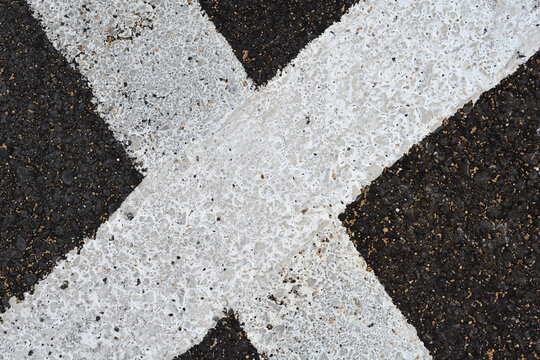 A close up top down view of thick weathered white painted lines crossing on a dark textured asphalt road surface.