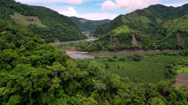 A scenic, high-angle drone establishing shot capturing the massive Grande de T&eacute;rraba river as it snakes through lush green valleys, dense rainforest canopies, and agricultural fields under a bright, p