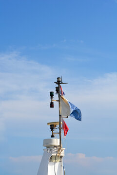 The white mast of a ship is equipped with a radar dome navigation lights and several fluttering flags against a clear blue sky