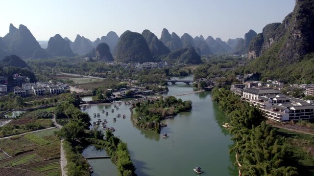 A scenic landscape of a winding river flanked by karst mountains, small settlements, boats, and farmland under clear skies.