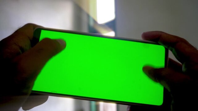 A person holding and playing on a smartphone with a green screen display, capturing mobile gaming, application use, and digital technology concepts in a horizontal view.