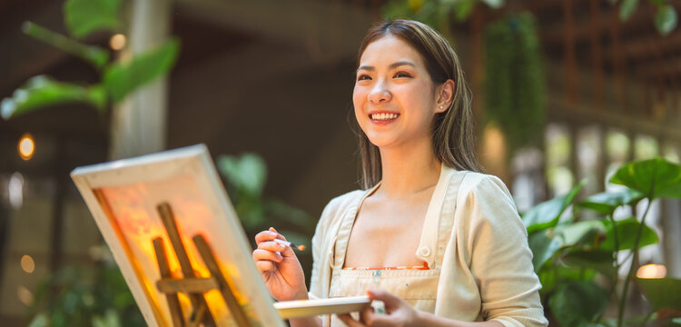 Portrait of happy young adult asian woman painting brush on canvas at workshop art lesson class.