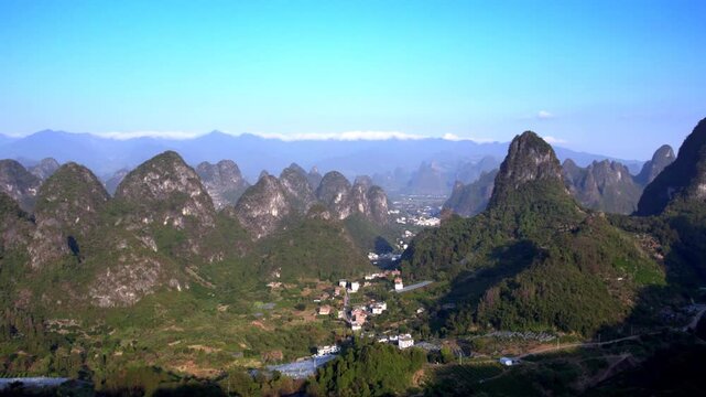 A panoramic landscape of steep limestone karst peaks framing a small settlement in a green valley under a clear blue sky.