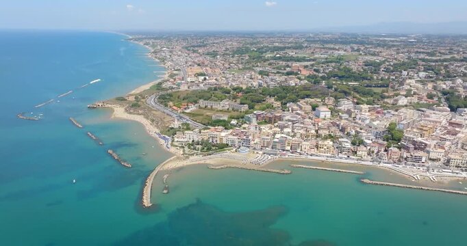 Panorama of Anzio seafront, located near Rome, Italy. Aerial view of a coastal town stretching along turquoise sea waters, sandy beaches, breakwaters, and urban buildings under a clear blue sky.