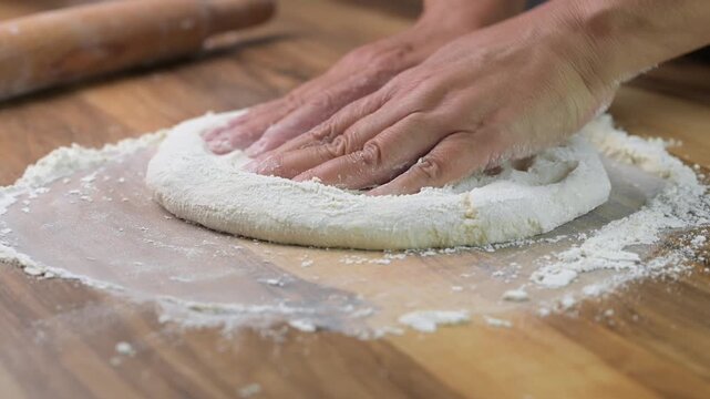 Close up baker hands kneading fresh pizza dough on wooden board with flour, traditional Italian cooking in pizzeria kitchen
