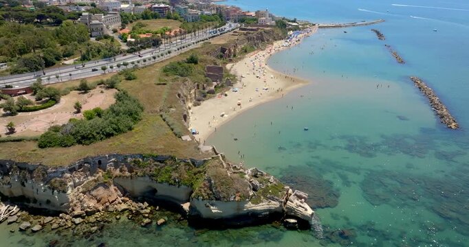 Aerial view of a scenic coastal beach with sandy shores, clear blue water, rocky cliffs, and a protective breakwater pier under a bright sunny sky. Panorama of Grotte di Nerone Beach in Anzio, Italy.