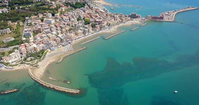 Aerial view of Anzio, near Rome, Italy, showing the historic port, breakwaters protecting the harbor, sandy beaches with sun umbrellas, and the city's waterfront buildings along the Tyrrhenian Sea.