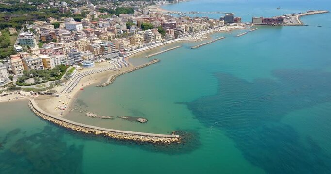 Aerial view of Anzio, near Rome, Italy, showing the historic port, breakwaters protecting the harbor, sandy beaches with sun umbrellas, and the city's waterfront buildings along the Tyrrhenian Sea.