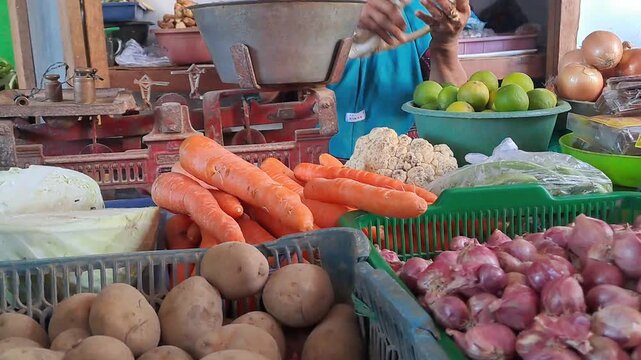 Indonesian market vendor weighing vegetables and spices for customer. Fresh shallots, garlic, chili, carrots, potatoes, limes displayed. Traditional market stall with vintage scale. Authentic local.