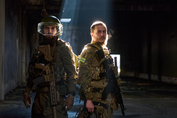 Obraz premium Two Soldiers in Tactical Gear Standing Back-to-Back Inside Dimly Lit Industrial Building