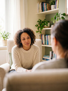 A female therapist with curly hair sits attentively in an armchair listening to a patient in a bright plant-filled consultation room.