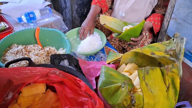 Indonesian vendor wrapping traditional ketan sticky rice in banana leaf. Served with cassava and grated coconut. Authentic jajanan pasar and traditional snack preparation in local market.
