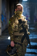 Obraz premium Masked Soldier In Camouflage Tactical Gear Holding Rifle And Helmet In Urban Setting