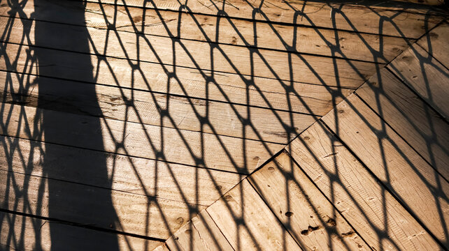 A close-up photograph featuring an intricate fence or net shadow cast over a warm, sunlit wooden deck surface, creating a geometric pattern and high-contrast textures.