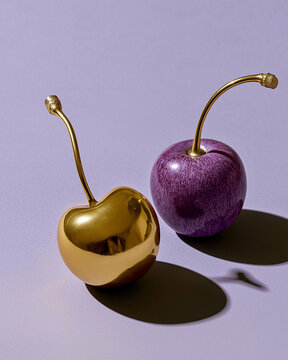 Two polished gold cherries with a purple cherry on a lavender surface. Ideal for luxury food, editorial, and premium branding.