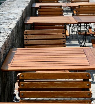 perspective view of a long row of empty wooden tables and chairs set up at an outdoor street cafe along a stone promenade wall with rocky mountains in the background. Ideal concept for restaurant