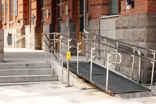 Accessible building entrance with non-slip wheelchair ramp, stainless steel handrails, and yellow assistance call button.