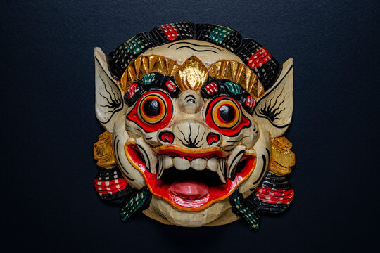 Traditional Balinese wooden demon mask on black background. Colorful carved wooden Barong or demon mask from Bali with big eyes and sharp teeth on dark black background, closeup, top view