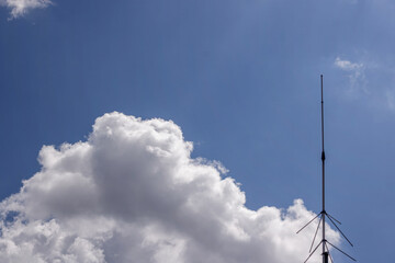 Obraz premium Vertical metallic antenna lightning rod mounted on a roof against a blue sky with fluffy white clouds in daylight. High Quality Photo