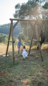 A Couple's Joyful Swing: A man gently pushes a woman on a wooden swing under a large olive tree, surrounded by a sun-drenched field. Their shared moment evokes warmth and idyllic happiness.