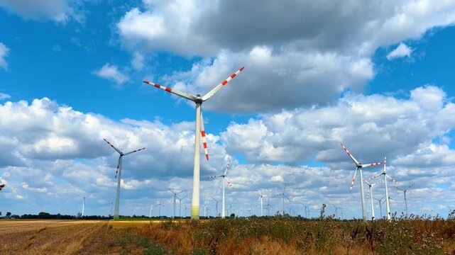 Clean energy wind farm in countryside under sunny sky. Tall electricity generators stand on agricultural land under white cumulus clouds in open landscape.