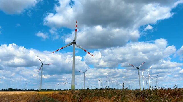 Wind turbines in rural field under blue sky with clouds. Tall power generators stand on agricultural land under sunlight with dry grass in foreground.