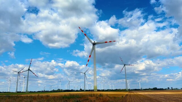 Alternative energy wind turbines in flat farm landscape. Modern electricity generators stand on dry golden field under bright sunlight and fluffy clouds.
