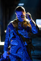 Naklejka premium Armed Soldier in Tactical Camouflage Gear Adjusting Face Mask While Holding Assault Rifle in Low Light