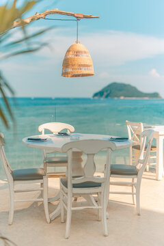 Charming seaside taverna dining table under hanging lamp beside ocean island view Mediterranean summer. Stylish coastal lifestyle hospitality scene. Romantic restaurant holiday, tranquil Greek getaway