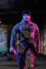 Obraz premium Armed Soldier In Camouflage Tactical Gear Holding Helmet And Assault Rifle In Urban Setting