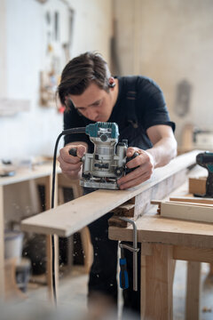 Concentrated master carpenter using handheld wood router instrument in restoration workshop, man restorer working with electric plunge router cutting off layers from board surface in repair studio.