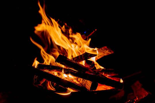 Burning campfire flames and firewood glowing against a black background, warm bonfire close-up