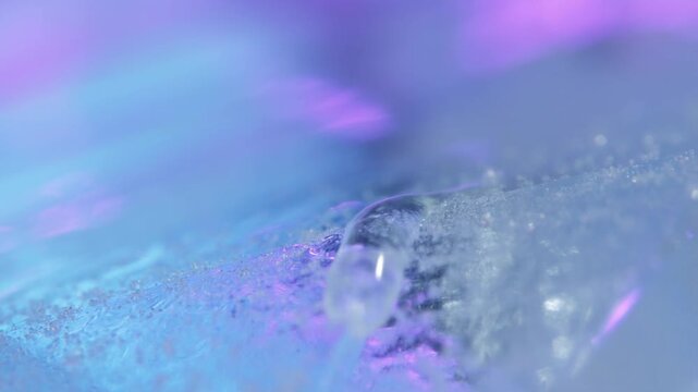 Slow motion macro tracking shot of water droplet sliding across icy textured surface with colored lighting