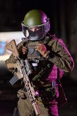 Fototapeta premium Armored Soldier In Tactical Gear Holding Assault Rifle With Riot Helmet And Ballistic Visor At Night