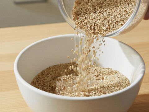 Mixed ancient grains pour from glass measuring cup into white bowl on wooden counter