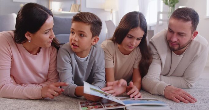 Happy family lying on carpet, enjoying quality time together while flipping through family photo album. Smiling parents and their children discussing photographs, sharing stories and good memories.